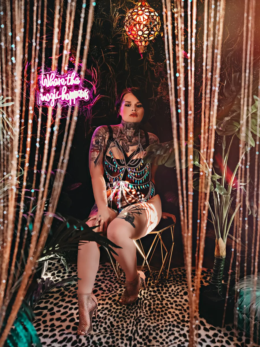 Tattooed client seated between beaded curtains on a leopard rug, under a pink neon sign